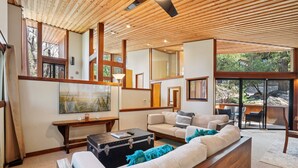 A modern living room with a wooden ceiling, large windows, a sofa, a coffee table, and a fireplace. - Idyllwild Realty Vacations (Idyllwild)