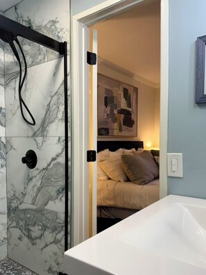 A bathroom with a white bathtub, a shower with marble tiles, and a bed with beige bedding in the adjacent room.