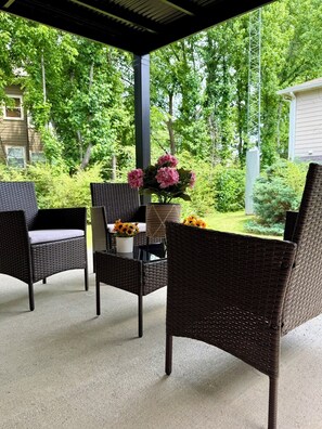 An outdoor patio with wicker furniture, a table with a vase of flowers, and a view of a green landscape.