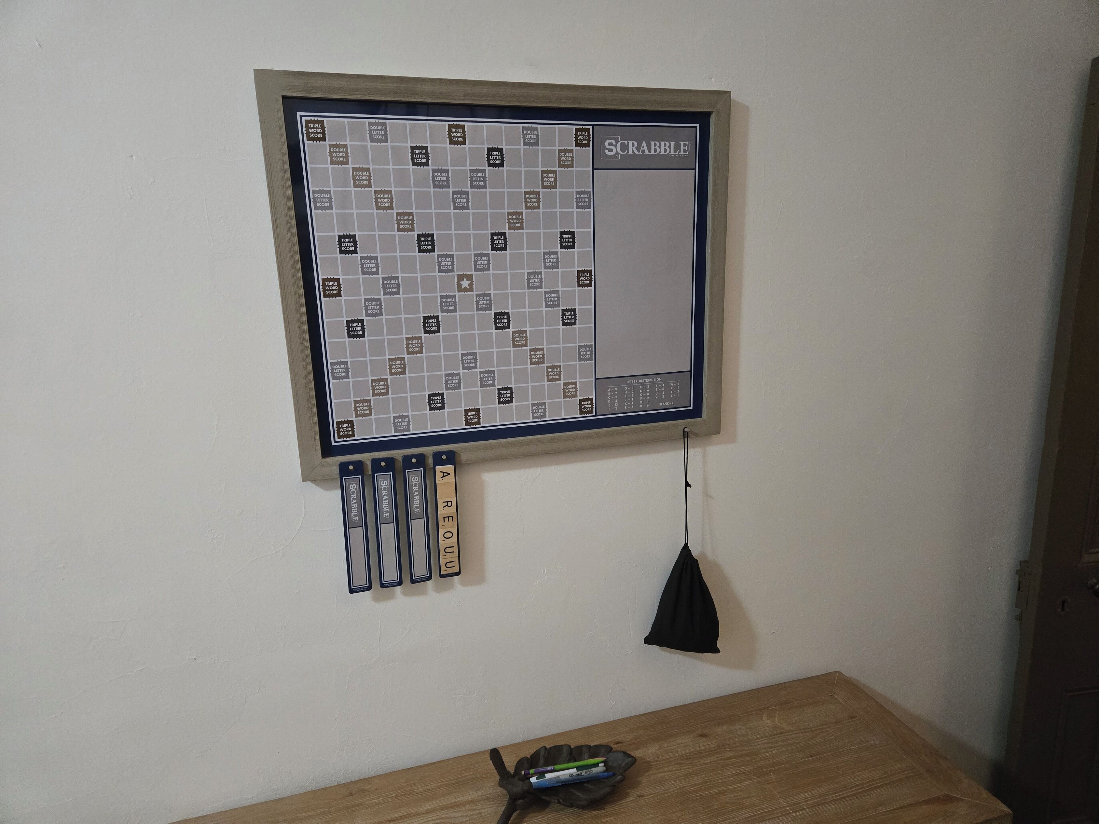 Ein Scrabble-Spiel auf einem Regal angebracht, mit abgehängten Buchstabenkacheln und einer schwarzen Fransen, die vom Brett baumelt.