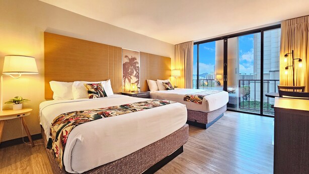 Een hotelkamer met twee bedden, een groot raam met uitzicht over de stad, en een tafeltje met een lamp.