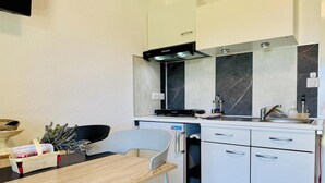 Une cuisine compacte équipée d’un four à micro-ondes, d’une cuisinière et d’un évier. On y trouve également un petit réfrigérateur, une table à manger avec des chaises, et un panier contenant divers objets posé sur la table.