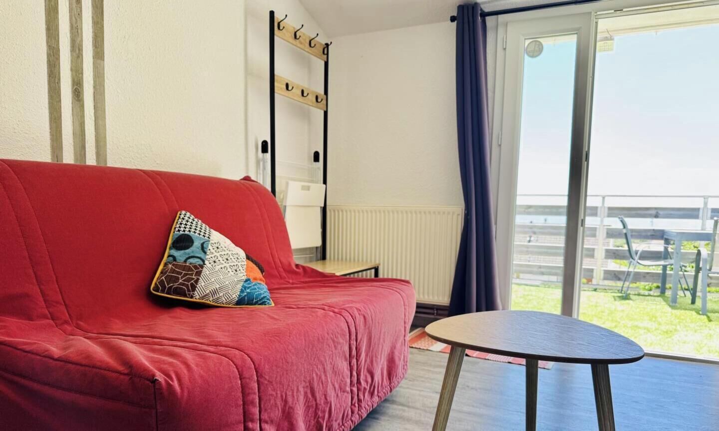 Een rode bank met een gekleurd kussen, een kleine tafel en uitzicht op een balkon met een hekwerk.