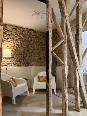 Una habitación con pared de piedra, dos sillas blancas con cojines, una lámpara de pie y vigas de madera.
