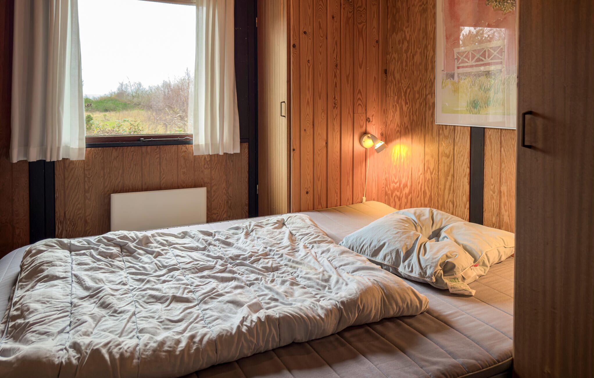 A bedroom with a wooden headboard, a bed with a white duvet and pillows, a window with curtains, and a wall-mounted light fixture.