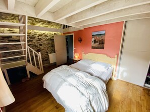 A bedroom with a bunk bed, a wooden staircase, a stone wall, a nightstand with a lamp, and a painting on the wall.