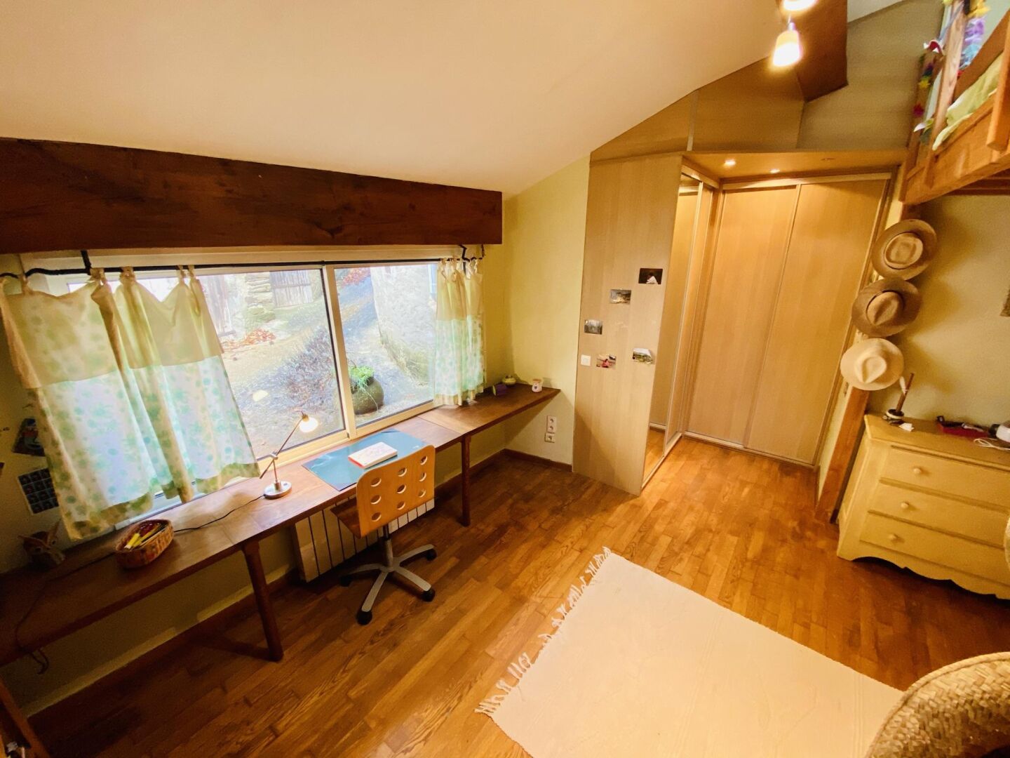 Een kamer met een houten bureau, een stoel, een raam met gordijnen, een deur en een wand met hoeden.