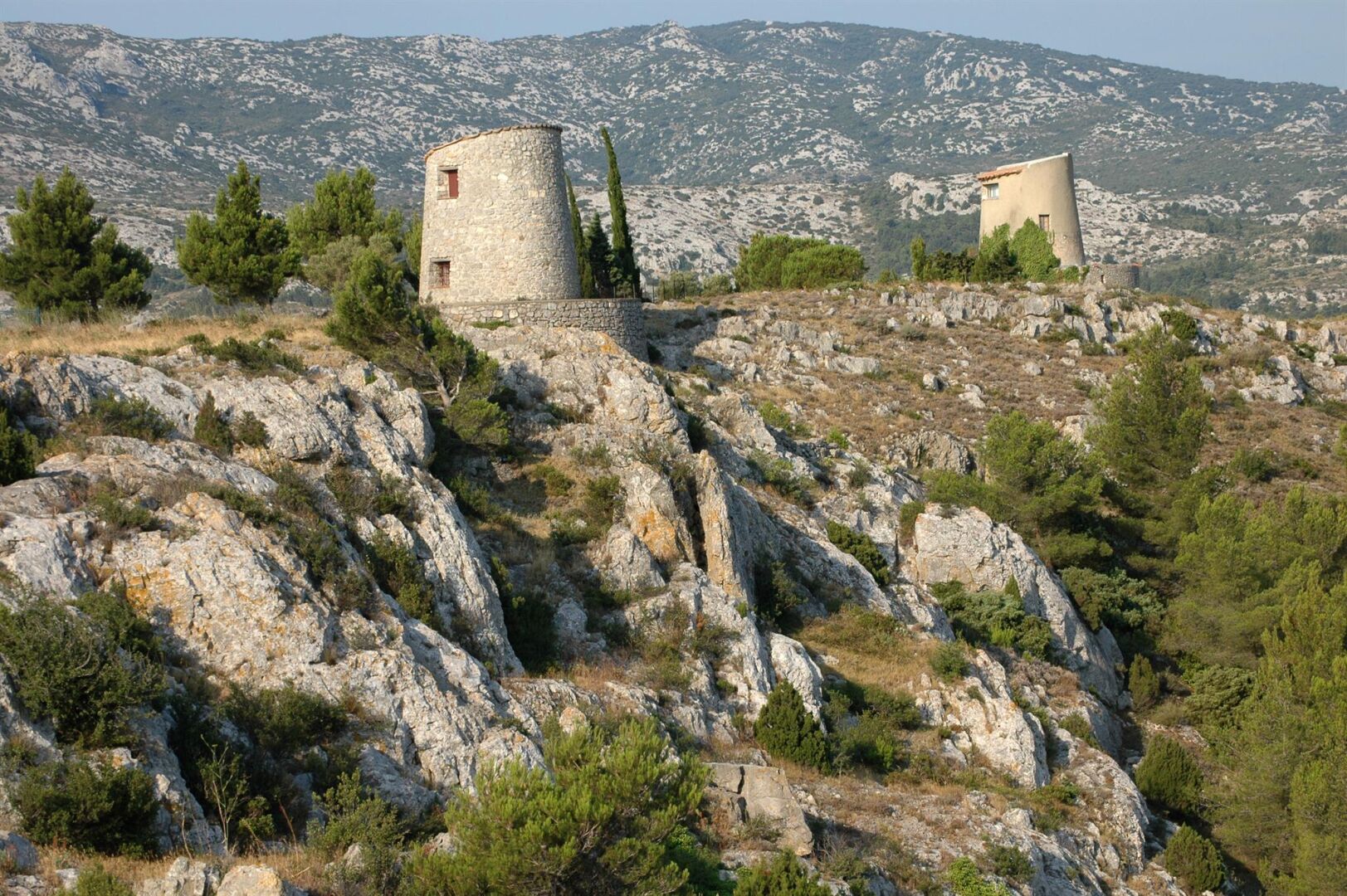 A mountainous landscape with two stone buildings, one cylindrical and one rectangular, surrounded by rocky terrain and sparse vegetation.