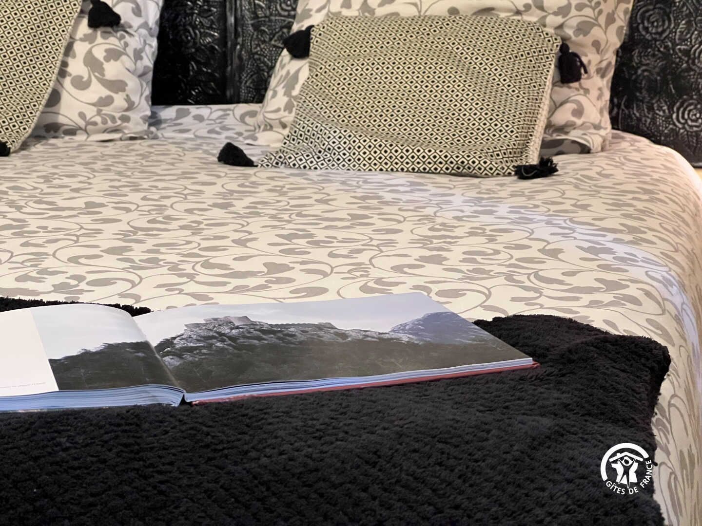 Una cama con ropa de cama estampada, un plaid negro y un libro con una imagen de montaña.