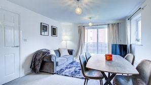 Ein Essbereich mit Tisch und Stühlen, einer Couch mit Decke, einer Lampe und einem Fenster mit Vorhängen.