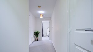 Ein schmaler Korridor mit weißen Wänden und einem Teppichboden. Links steht ein Blumentopf, rechts eine Tür.