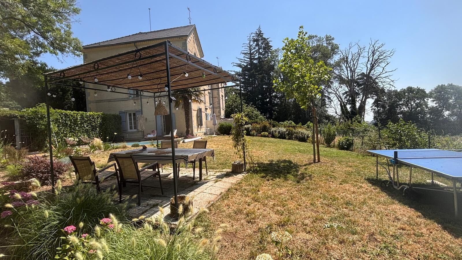 Un espacio exterior residencial con un porche cubierto, una mesa con sillas, una mesa de ping pong y una piscina.