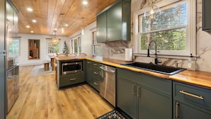 A modern kitchen with wooden ceiling, green cabinets, a stainless steel sink, and a large window with a view of trees.
