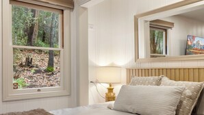 A bedroom with a bed, a window with a forest view, a bedside lamp, and a mirror on the wall.