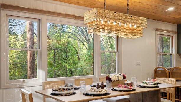 A dining area with a long wooden table set for a meal, large windows with a forest view, and a hanging light fixture.