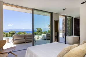 Una camera da letto moderna con un letto grande, una porta vetrata che dà su un balcone con vista sul mare e sulle montagne, e un bagno separato con una vasca.
