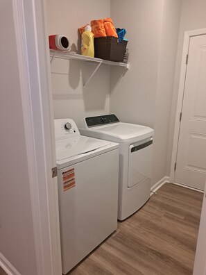 Ein Waschraum mit einer Frontlader-Waschmaschine und einem Trockner, ein Regal mit Waschmitteln und einer Tür zu einem anderen Raum.