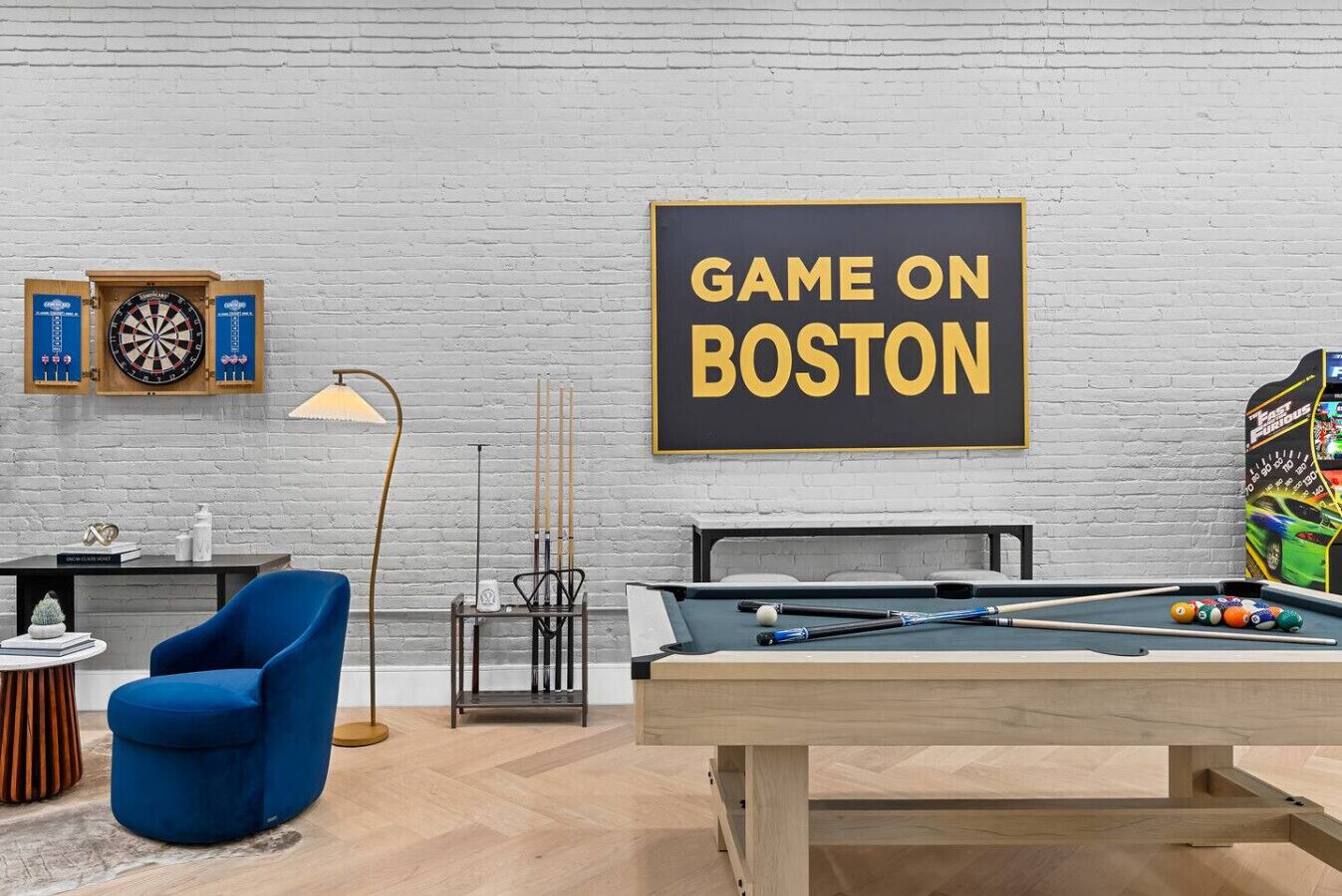 A room with a pool table, a dartboard, a blue chair, and a wall sign that reads "GAME ON BOSTON".