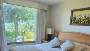 A neatly made bed with a beige blanket and pillows, a bedside table with a lamp, and a window with curtains. - Bordelago Pucón Playa Privada (Araucanía)