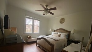 Un dormitorio con cama, mesitas de noche, escritorio, silla, ventilador de techo, ventana y radiador.
