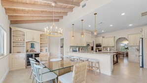 A spacious kitchen with white cabinetry, a central island, and a dining area with glass-top table and wicker chairs. - Waterfront Lux 4BR! New Reno with Heated Pool + Outdoor Kitchen (Lighthouse Point)