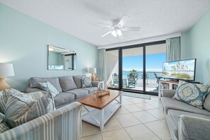 Uma sala de estar moderna com um sofá, uma mesa de centro e uma televisão. Há uma janela grande com vista para o oceano.
