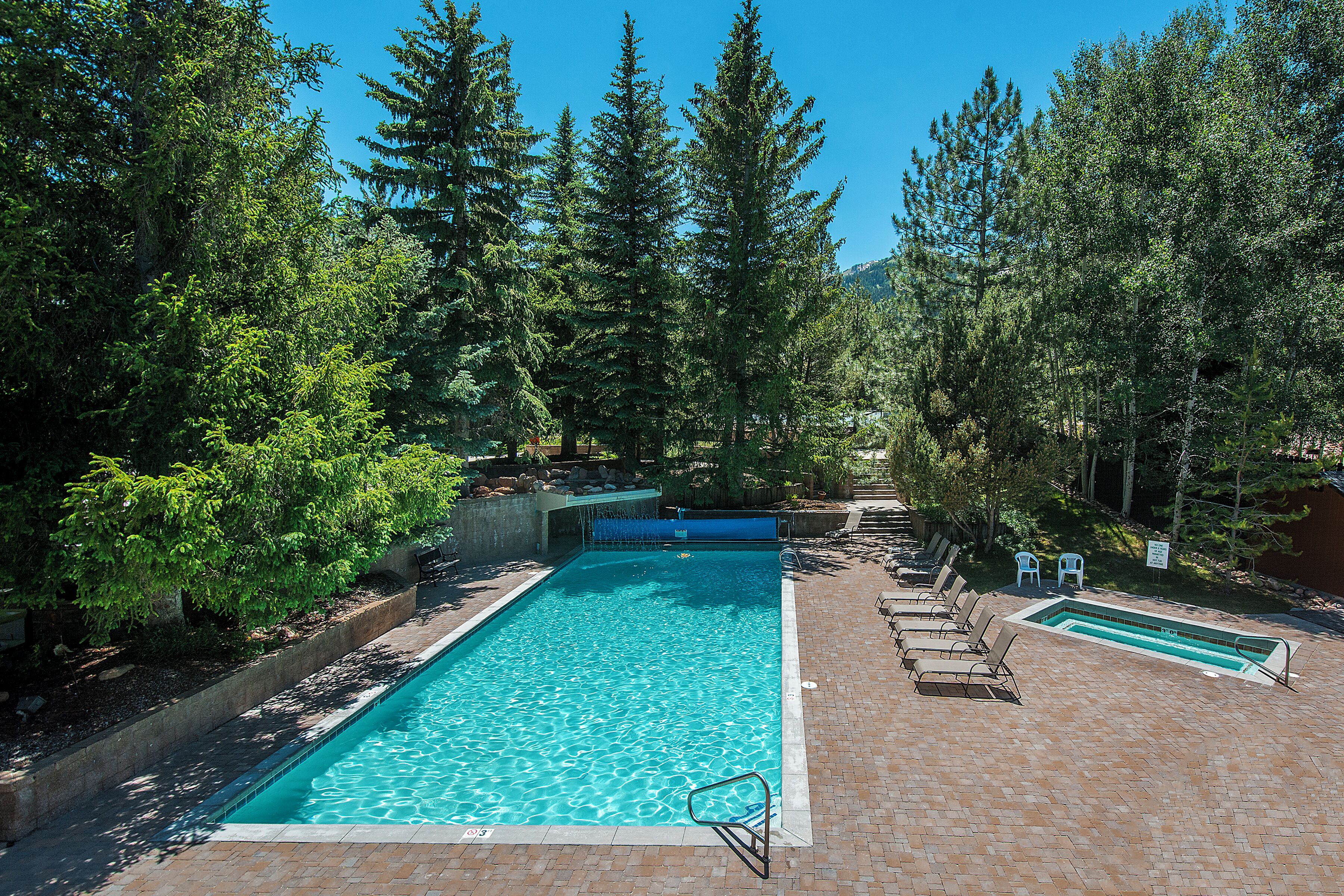 A rectangular swimming pool with a clear blue water surface, surrounded by a brick patio and wooden lounge chairs.