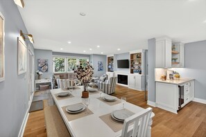 Une cuisine moderne avec des meubles blancs, un coin repas avec une longue table dressée pour six personnes, et un salon avec un canapé et une télévision.
