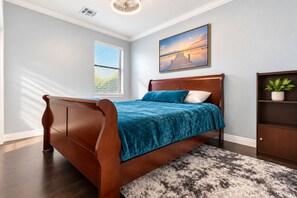 A bedroom with a wooden bed, a blue bedspread, a white pillow, a dark wooden nightstand, a potted plant, and a painting of a sunset on the wall. - Perfect 1-level, 4-bedroom, 2100 sqft house with AC in charming Avondale, AZ (Avondale)