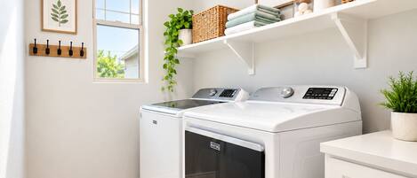 A laundry room with a front-loading washer and dryer, a shelf with baskets and a framed sign, and a window with a plant.