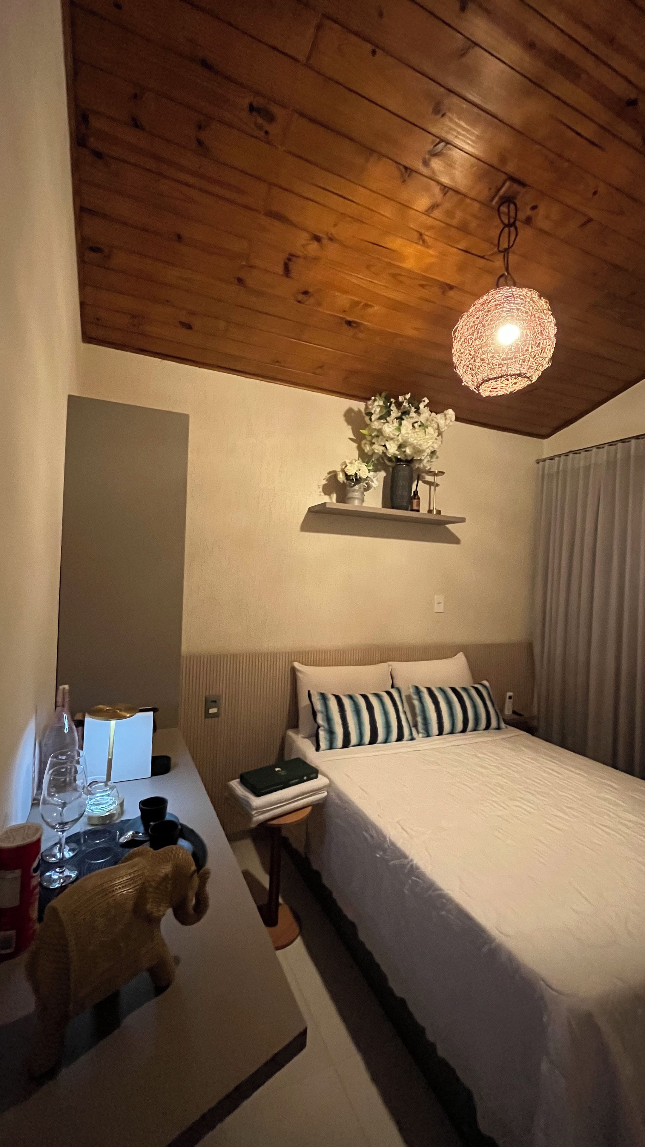 A bedroom with a wooden ceiling, a bed with striped pillows, a nightstand with a lamp, and a hanging light fixture.