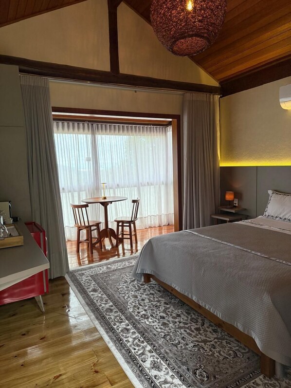 Un dormitorio con una cama grande, una mesa de comedor con sillas, una mesita de noche con lámpara y vistas al exterior a través de un ventanal amplio.