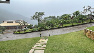 A paved pathway leading to a building with a sloped roof, surrounded by greenery and a stone wall.