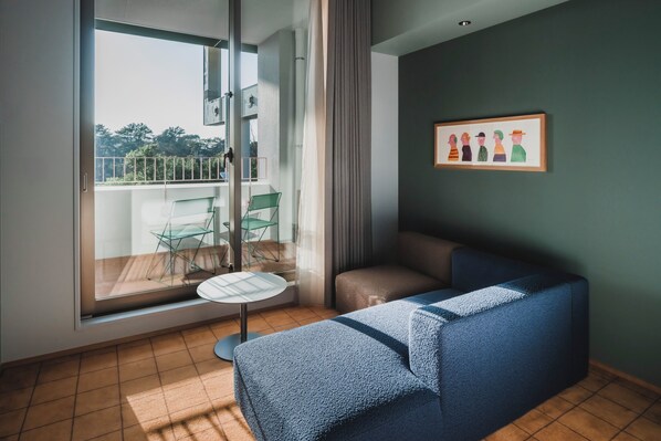 A modern living room with a blue sofa, a small round table, and a glass door leading to a balcony. - THE TSUBAKIMORI Hotel & Residence (Chiba)