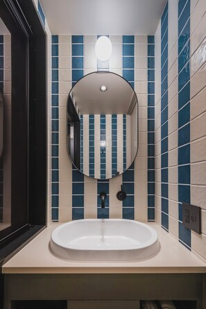 A bathroom with a round mirror, a sink, and blue and beige tiled walls. - THE TSUBAKIMORI Hotel & Residence (Chiba)