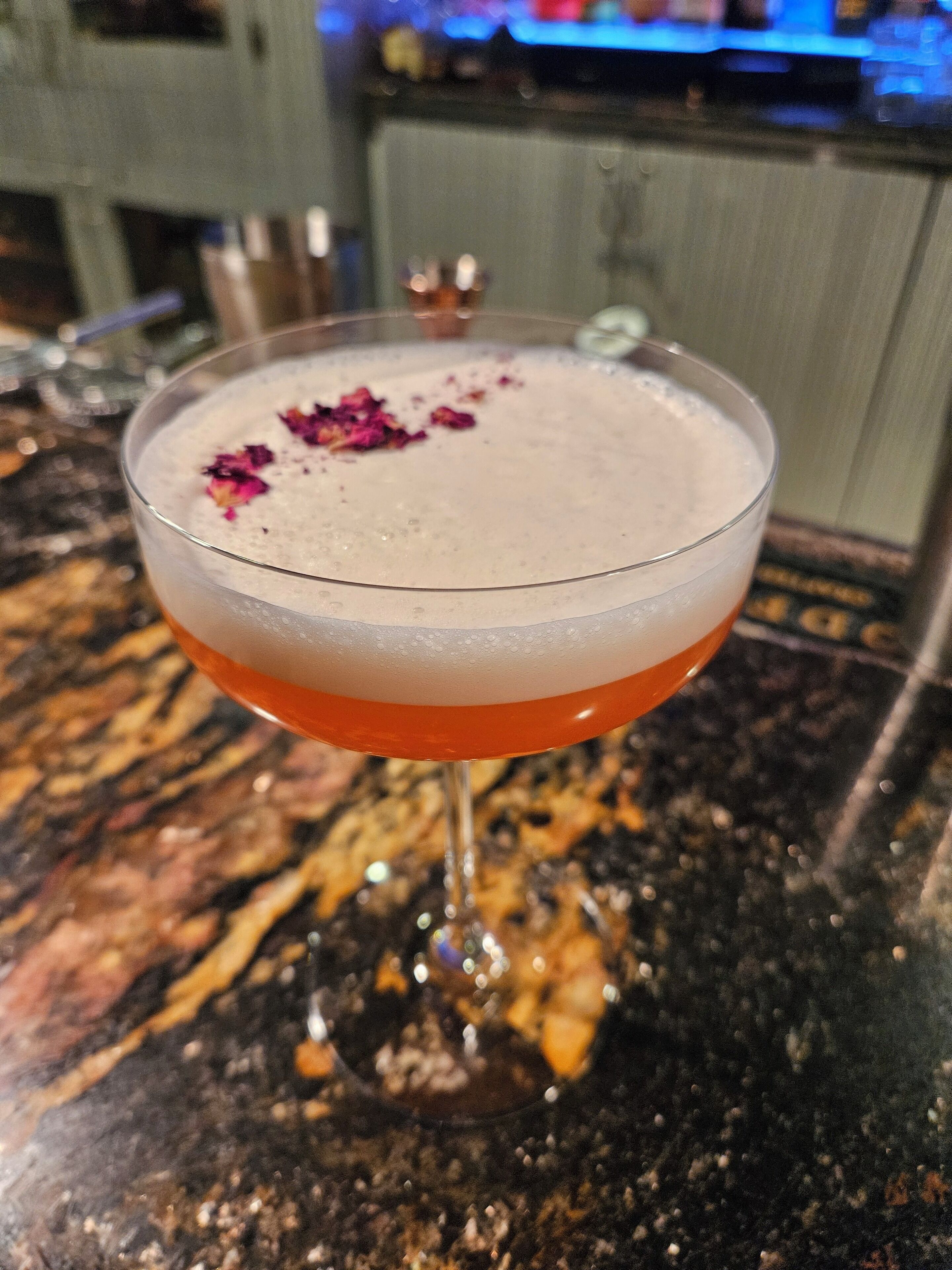 En cocktail i et martiniglas med skummende top, pyntet med lyserøde blade, på en marmor bordplade.