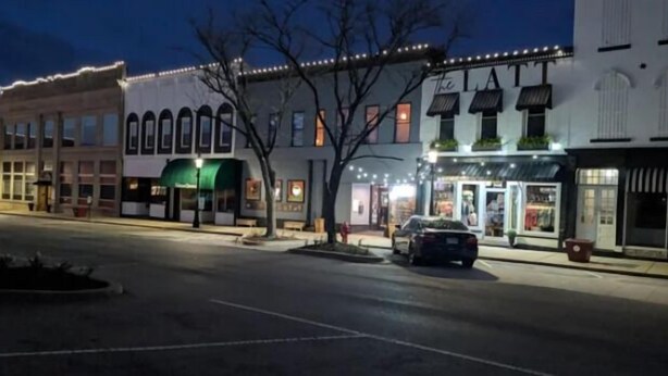A street view at night with buildings, a car parked on the side, and a tree in the foreground.