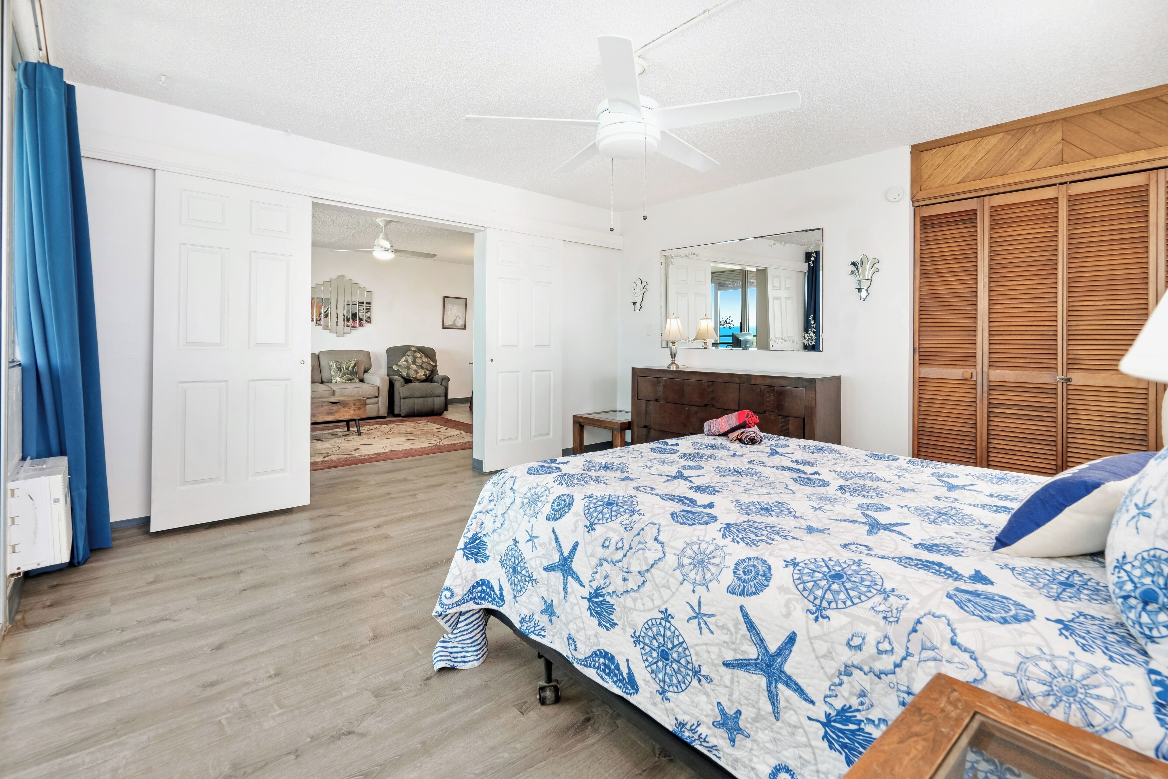 A bedroom with a bed, a wooden wardrobe, a ceiling fan, and a view into another room.