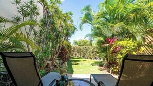 Une terrasse avec deux chaises et une table en verre, entourée d’une végétation tropicale luxuriante et d’une pelouse bien entretenue.