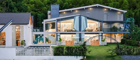 Ein modernes, zweistöckiges Haus mit großer Glasfassade, einer sichtbaren Außenlounge und einem gepflegten Garten.