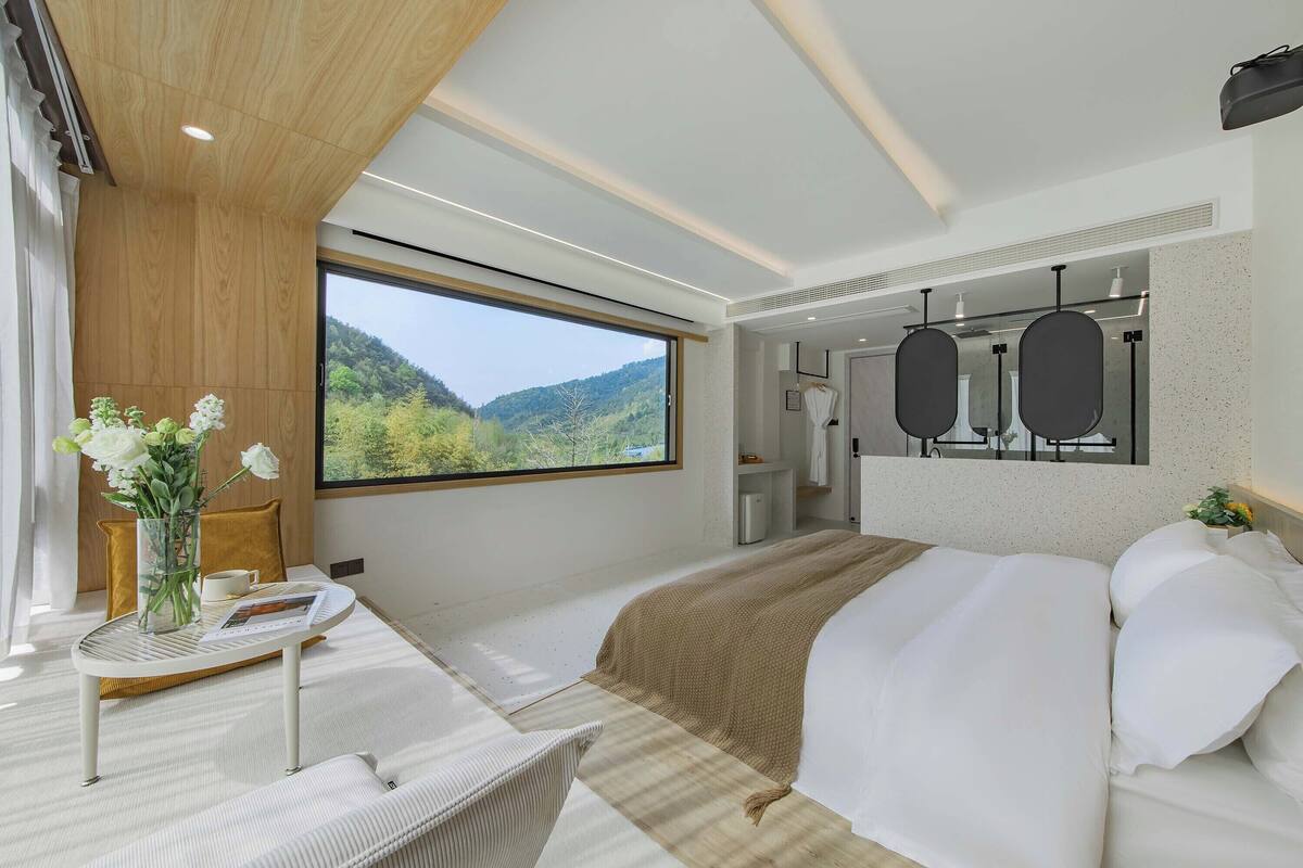 Ett modernt hotellrum med en stor säng, ett skrivbord med en stol, ett fönster med utsikt över bergen och ett badrum med en spegel och handduksställ.