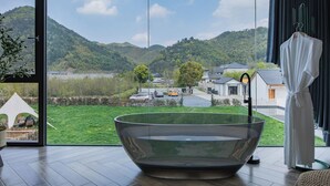 A modern room with a large glass bathtub, a view of a green landscape, and a white robe hanging by the window. - Qingcen Manor (Huzhou)