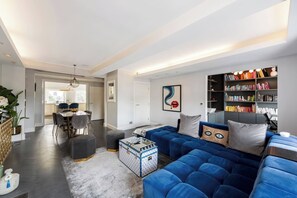 A modern living room with a blue sofa, a dining area, and a bookshelf filled with books. - 5-bed Penthouse Huge Roof Terrace AC (London)