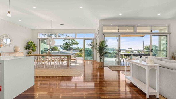 A spacious living area with a wooden floor, a dining table with chairs, a sofa, and a large sliding glass door leading to a balcony with a view of the sea. - Aerial Blue Mollymook - Ocean Views, Spacious, Wood Fireplace, Walk to Mollymook Beach & Golf Course (Mollymook)