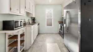 Una cocina con armarios blancos, un refrigerador de acero inoxidable, un microondas y una ventana con persianas.
