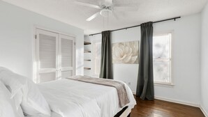Un dormitorio con cama, paredes blancas, ventilador de techo y ventana con cortinas.