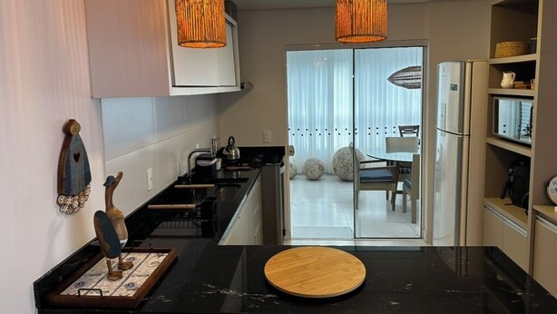 Una cocina moderna con encimeras negras, dos lámparas colgantes naranjas, una tabla de cortar de madera y vistas al área de comedor a través de puertas de vidrio.