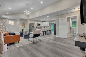 A modern living room with a sofa, armchair, and coffee table, a kitchen area with cabinets and appliances, and a dining area with chairs and a table.