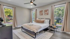 Une chambre moderne comprenant un lit, un bureau, une chaise, un ventilateur de plafond et de grandes fenêtres avec des rideaux.