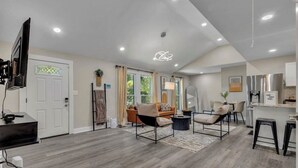 Un salon moderne avec un canapé, des fauteuils et une table basse. Un téléviseur est fixé au mur, il y a une porte avec un panneau de verre et un luminaire au plafond.